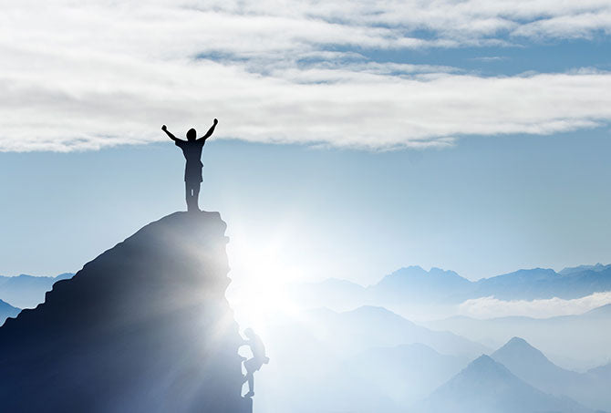 Symbolbild – Mann steht in Siegerpose auf Gipfel, weil er wichtige Aufgaben mit Erfolgsjournal erledigt hat